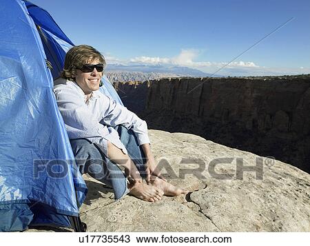 Stock Image - Young man sitting in tent by cliff, Moab, Utah, USA. Fotosearch