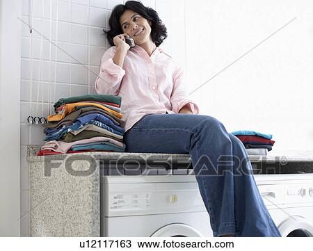 Stock Image - Mid adult woman sitting on washing machine and talking on phone. Fotosearch