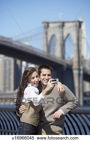 Stock Photography - Couple taking photograph with Brooklyn Bridge in background, New York City, New York, USA. Fotosearch