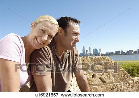 Stock Photo - Mature couple sitting by brick wall, skyline in background. Fotosearch