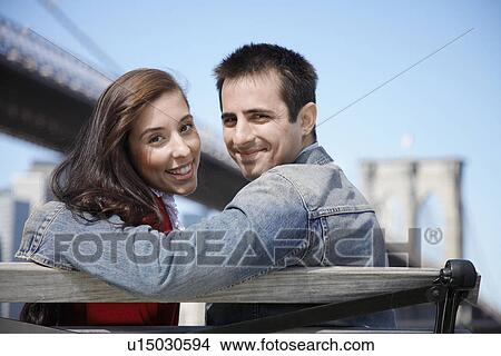 Picture - Young couple sitting on bench, looking over shoulders (rear view), Brooklyn Bridge, New York City, New York, USA. Fotosearch