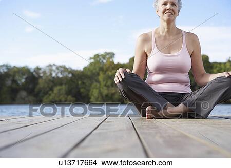 Picture - Mature woman sitting crossed legged by lake (low angle view). Fotosearch