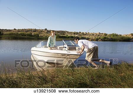 Stock Photo - Young man pushing motorboat to lake, girlfriend looking. Fotosearch