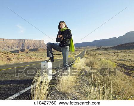 Stock Image - Female hitchhiker checking time on rural road. Fotosearch