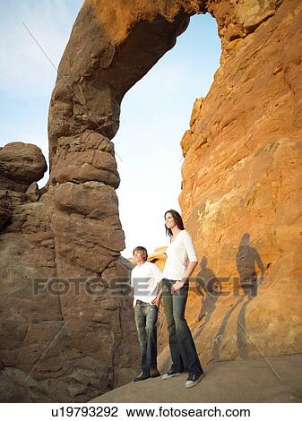 Stock Image - Man and woman standing by arched rock, low angle view. Fotosearch