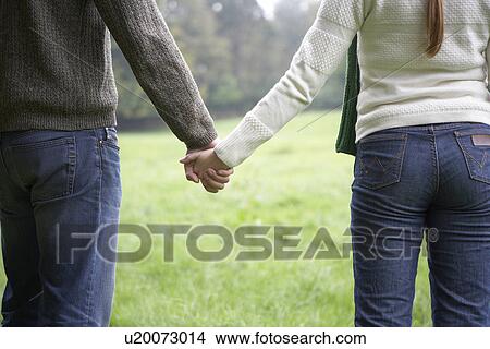 Picture - Mid adult couple holding hands in field (midsection, rear view). Fotosearch