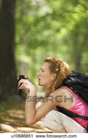 Stock Image - Woman crouching and using binoculars in forest. Fotosearch