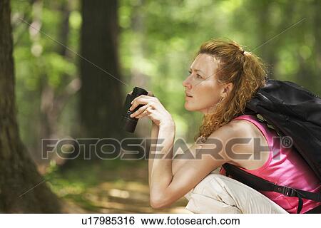 Stock Photograph - Woman crouching and using binoculars in forest. Fotosearch