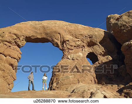 Stock Image - Young couple standing under arched rock. Fotosearch
