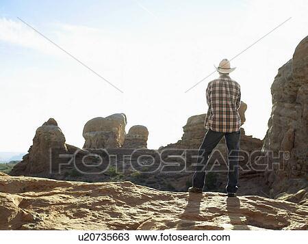 Stock Image - Young man in cowboy hat standing on cliff, rear view. Fotosearch