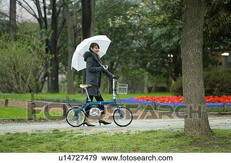 Stock Photo - Young Woman Pushing the Bicycle in the Park, Side View . Fotosearch