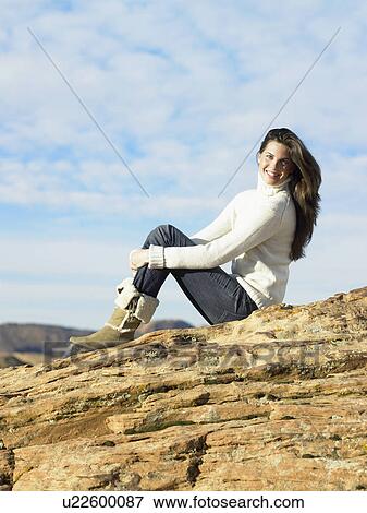 Stock Photo - Young woman sitting on rocky hill, portrait, low angle view. Fotosearch