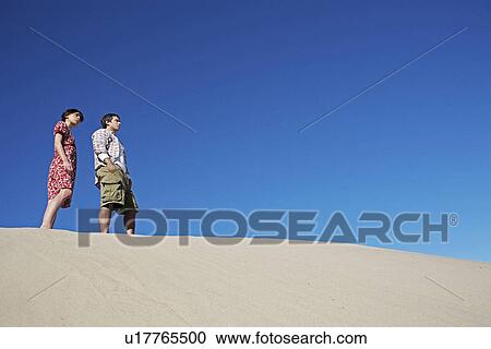 Stock Image - Couple Standing on Sand Dune (Low Angle View). Fotosearch