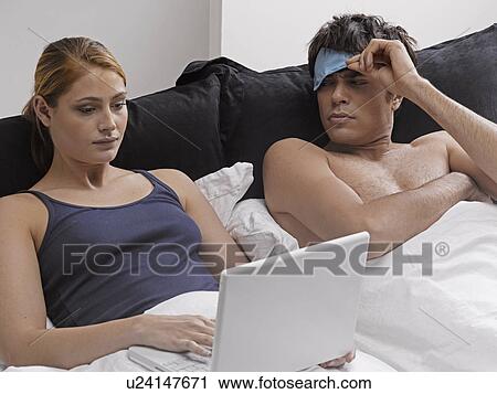 Stock Image - Young Woman Using Laptop in Bed, Frustrated Boyfriend Lying Beside Her. Fotosearch