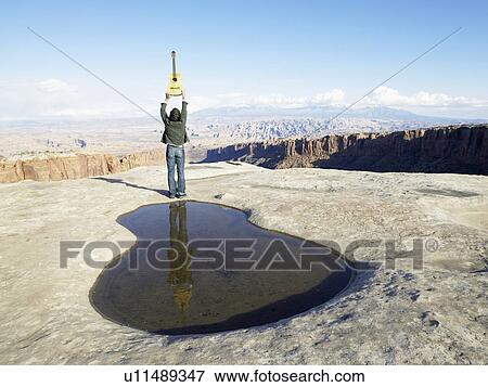 Stock Photo - Man holding guitar near puddle, rock strata in background, rear view, Moab, Utah, USA. Fotosearch