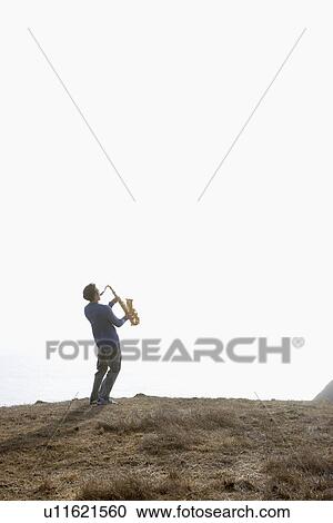 Stock Image - Man playing saxophone on cliff. Fotosearch