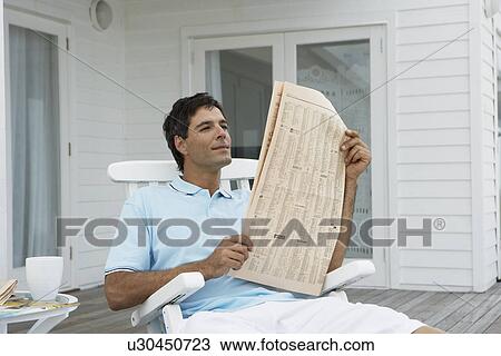 Stock Image - Mid adult man reading newspaper on veranda. Fotosearch