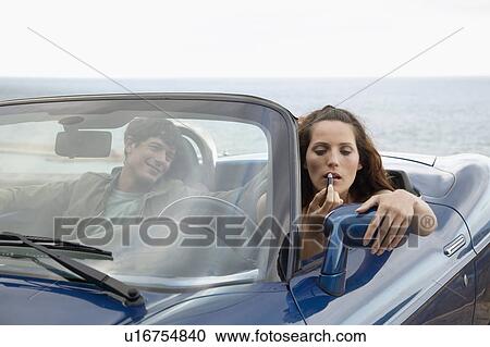 Stock Image - Woman Applying Lipstick in Convertible Beside Boyfriend. Fotosearch