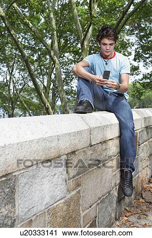 Stock Image - Teenage boy using mobile phone on stone fence. Fotosearch