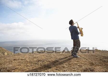 Stock Image - Man playing saxophone on cliff. Fotosearch