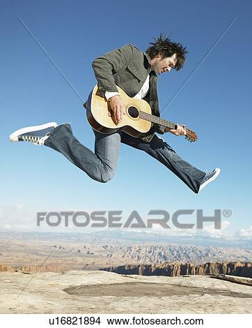 Picture - Mid adult man jumping and playing acoustic guitar, rock strata in background. Fotosearch