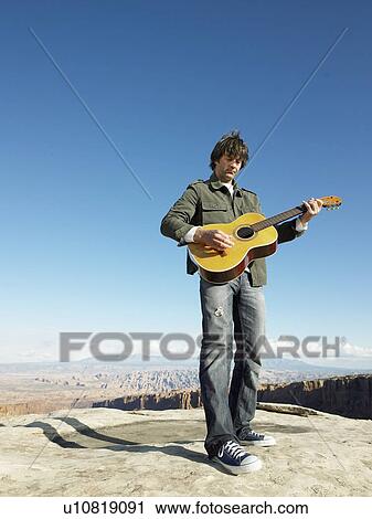 Stock Image - Mid adult man playing acoustic guitar, rock strata in background, low angle view. Fotosearch