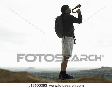 Stock Image - Mid adult man playing bugle on rock. Fotosearch