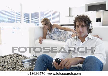 Stock Image - Young man listening to MP3 player, woman reading magazine on sofa. Fotosearch