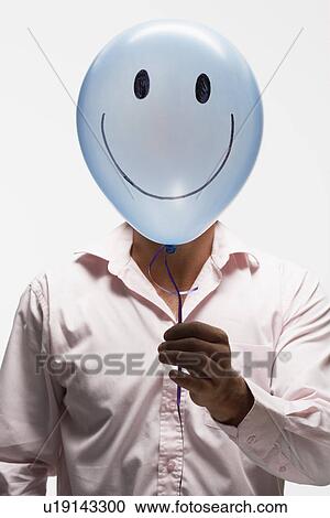 Stock Image - Man holding balloon with smiley face in front of his face. Fotosearch