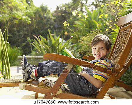 Stock Image - Boy reading book on garden chair. Fotosearch