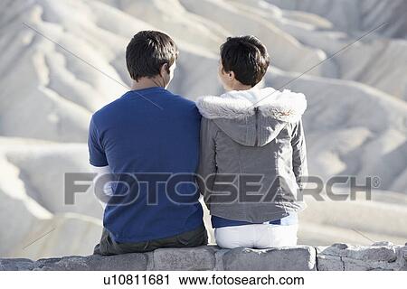 Stock Image - Couple sitting on stone wall in desert (rear view). Fotosearch
