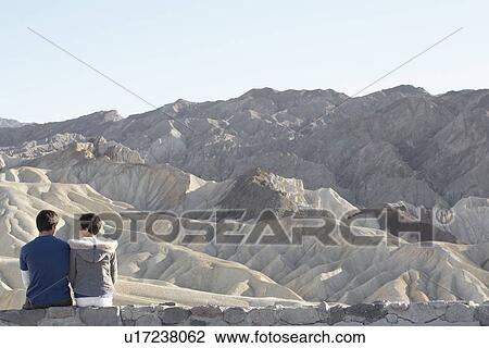 Stock Image - Couple sitting on stone wall in desert (rear view), Death Valley, California, USA. Fotosearch