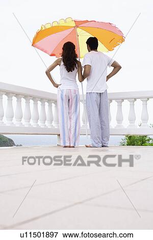 Stock Photo - Young couple standing under parasol and looking at view. Fotosearch