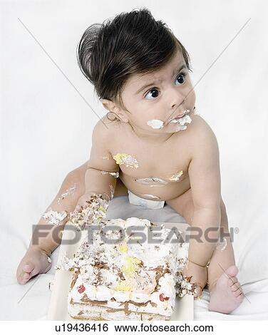 Stock Photograph - Close-up of a baby boy sitting in front of a cake with icing on his body. Fotosearch