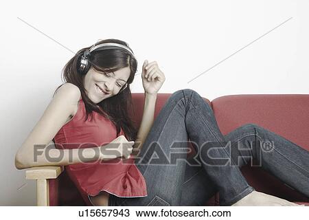 Young woman sitting on a couch wearing headphones and listening to music View Large Photo Image Stock Image - Young woman sitting on a couch wearing headphones and listening to music. Fotosearch