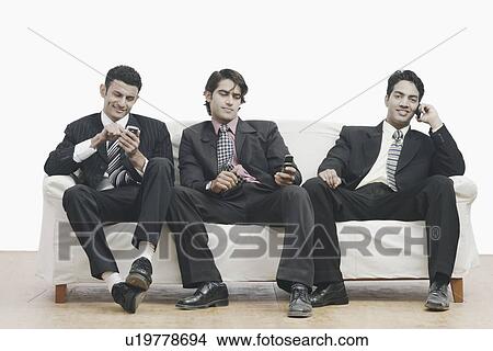 Three businessmen sitting on a couch holding mobile phones View Large Photo Image Picture - Three businessmen sitting on a couch holding mobile phones. Fotosearch