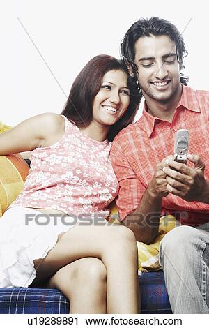 Stock Image - Young man holding a mobile phone with a young woman sitting on a couch. Fotosearch