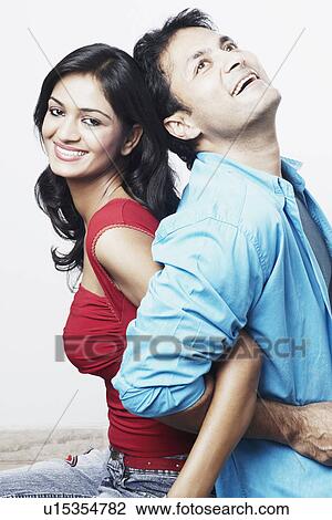 Stock Image - Side profile of a young couple sitting back to back and smiling. Fotosearch
