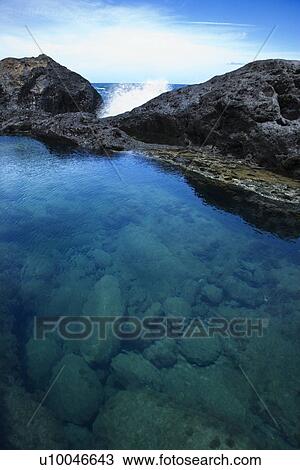 Stock Image - Tidal pool with wave crashing on rocks in background with blue sky in Maui, Hawaii, USA.. Fotosearch