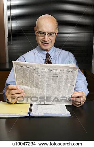 Picture - Caucasian middle-aged businessman sitting at desk in office reading newspaper smiling.. Fotosearch