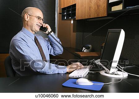 Caucasian middle-aged businessman sitting at desk in office talking on telephone and typing on computer. View Large Photo Image Stock Image - Caucasian middle-aged businessman sitting at desk in office talking on telephone and typing on computer.. Fotosearch