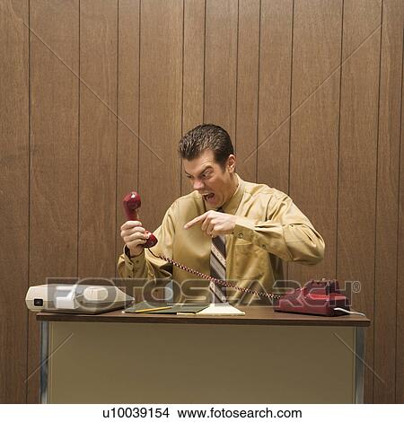 Picture - Caucasion mid-adult retro businessman sitting at desk pointing at telephone in anger.. Fotosearch