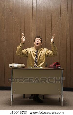 Stock Image - Caucasion mid-adult retro businessman sitting at desk raising hands in air with look of frustration.. Fotosearch