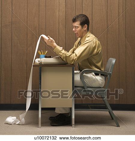 Stock Image - Caucasion mid-adult retro businessman sitting at desk looking with shocked expression at long adding machine printout that reaches all the way to the floor.. Fotosearch