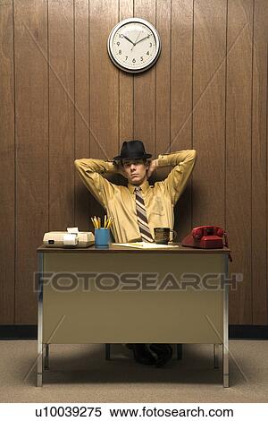 Stock Photography - Caucasion mid-adult retro businessman wearing fedora sitting at desk leaning back with hands behind head.. Fotosearch