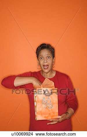 Picture - African American mature adult female with hand in gift bag looking surprised.. Fotosearch