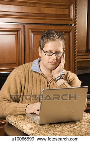 Stock Photo - Caucasian man working on laptop computer in kitchen.. Fotosearch