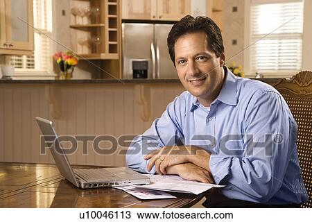 Stock Image - Caucasian mid-adult man paying bills on laptop computer looking at viewer and smiling.. Fotosearch