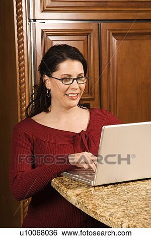 Stock Photograph - Caucasian woman typing on laptop in kitchen and smiling.. Fotosearch