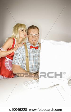 Stock Photo - Caucasian young blonde woman dressed in french maid outfit whispering to Caucasian young man sitting behind computer dressed like nerd.. Fotosearch